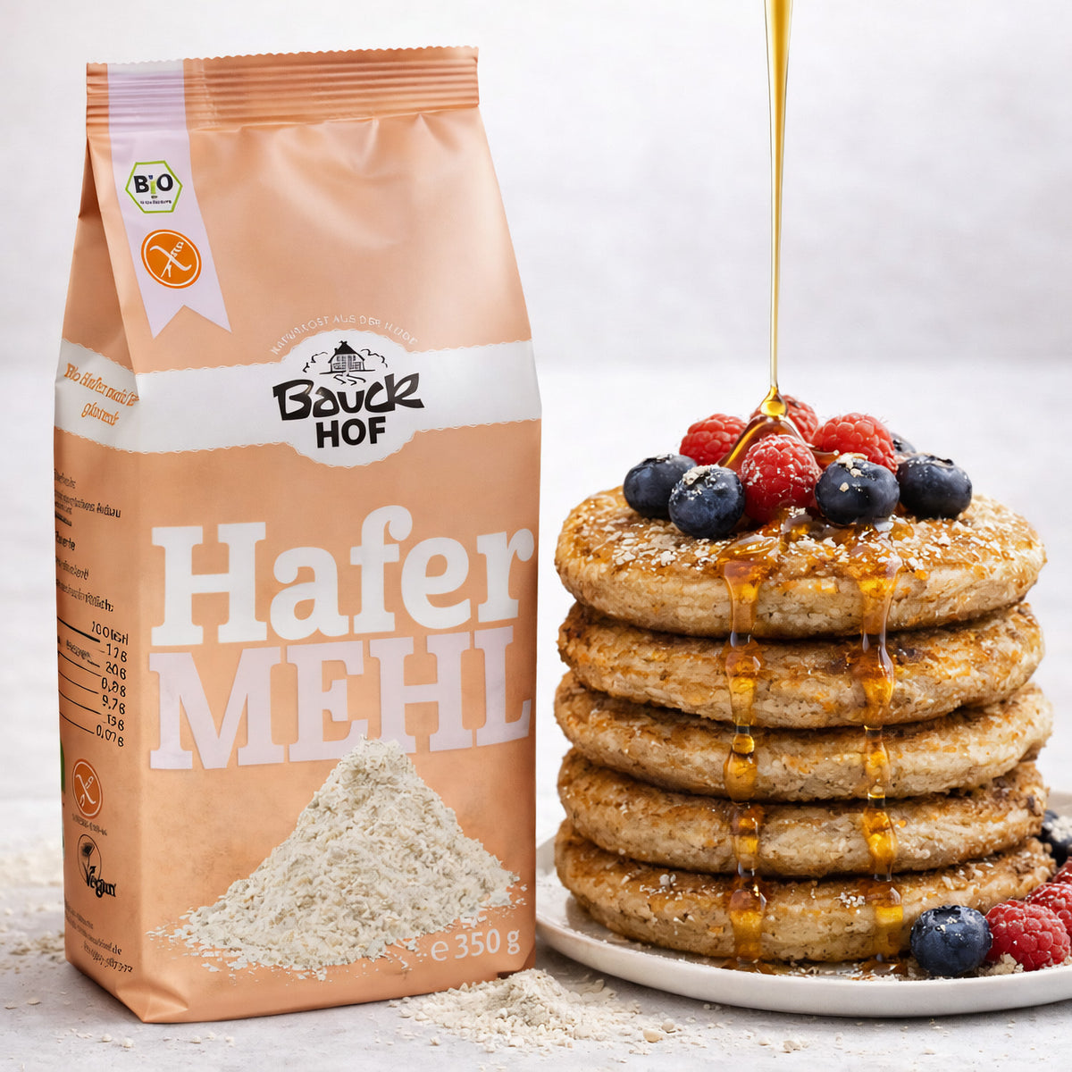 Packung bauckhof hafermehl vollkorn glutenfrei neben gestapelten Pancakes mit Beeren und Sirup, Hafermehl auf dem Tisch verstreut.