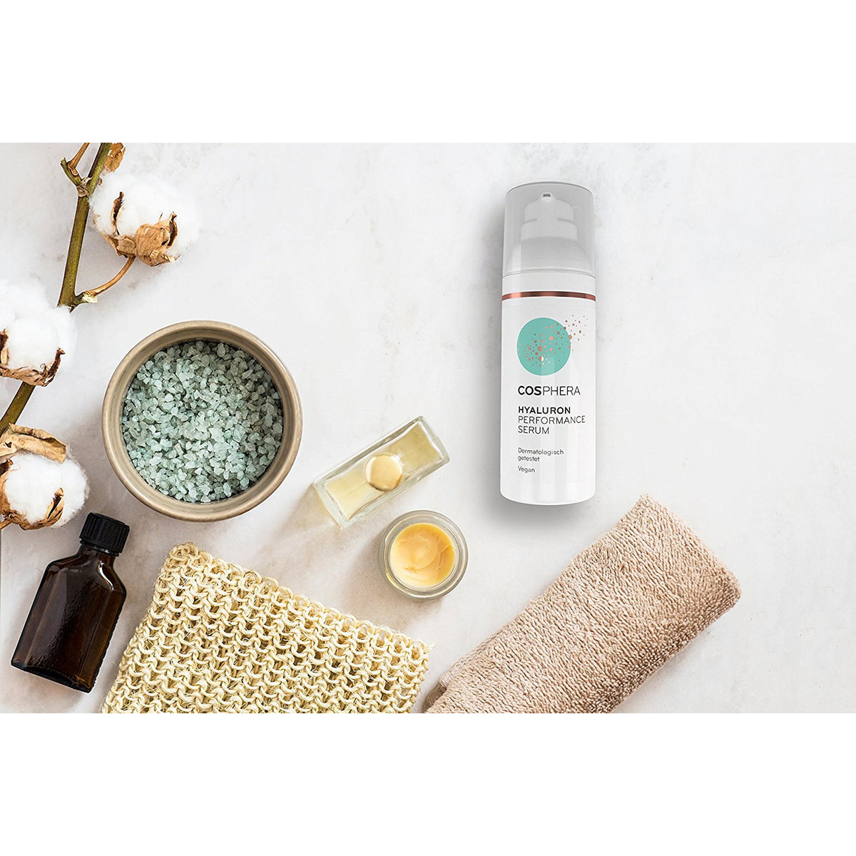 Cosphera Hyaluron Performance Serum Flasche auf weißem Untergrund, umgeben von Pflegeutensilien wie Salz, Öl, Creme und Baumwolle.