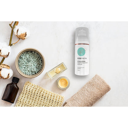 Cosphera Hyaluron Performance Serum Flasche auf weißem Untergrund, umgeben von Pflegeutensilien wie Salz, Öl, Creme und Baumwolle.