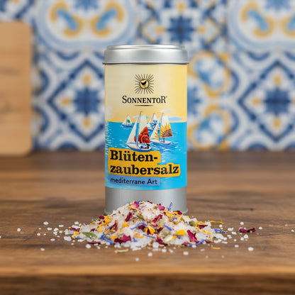 Capture a close-up shot of the 'Sonnentor Bio Blütenzaubersalz mediterrane Art' jar placed prominently on a rustic wooden kitchen counter illuminated by softbox lighting to highlight the vibrant floral and herb textures within the salt mixture. Use a macro