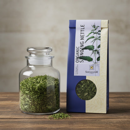 Capture a high-resolution close-up of Sonnentor Bio Brennnessel Tee lose in a clear, elegant glass jar, illuminated by softbox lighting that highlights the vibrant green of the tea leaves. The scene is set against a minimalist, neutral-colored background w
