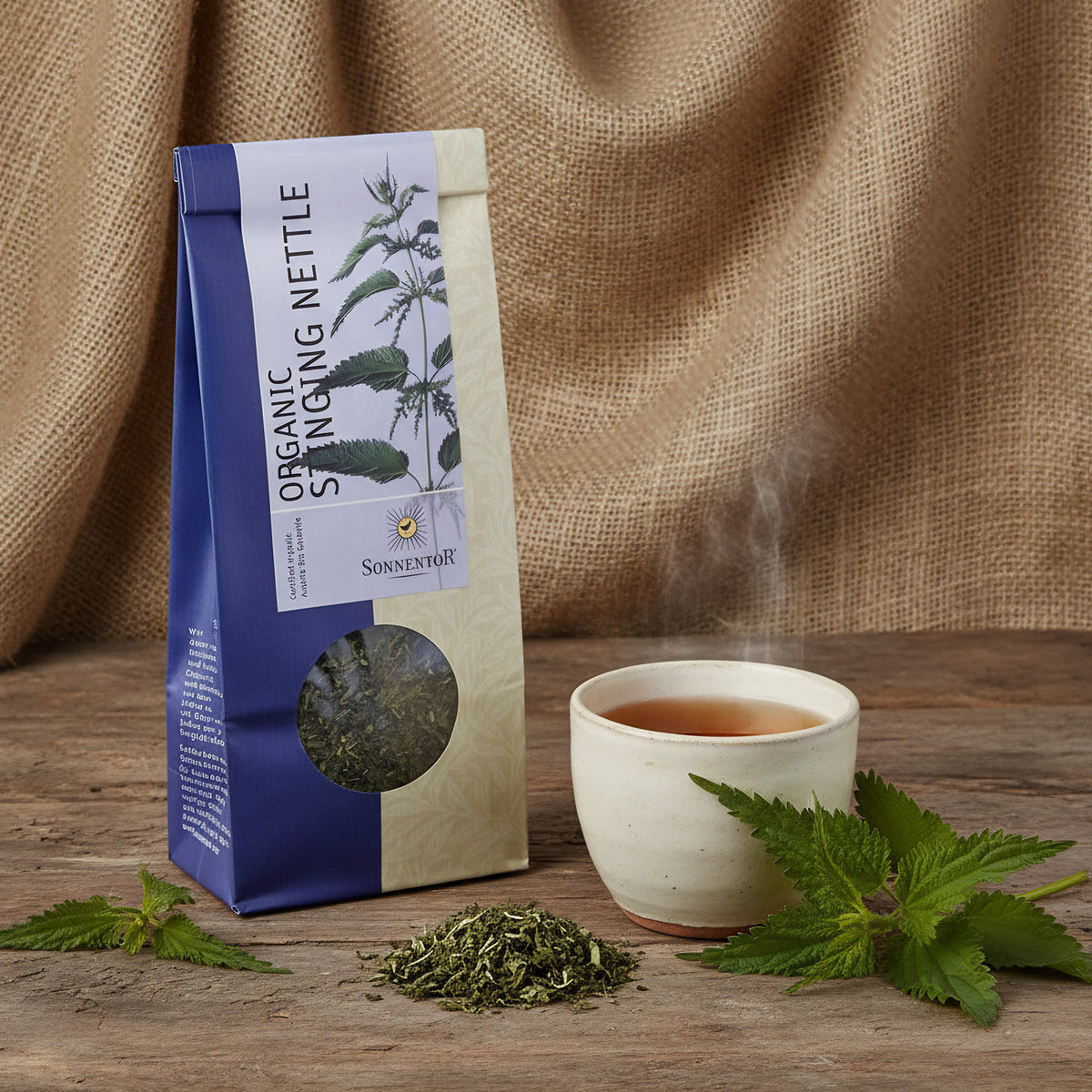 Create a studio shot of the Sonnentor Bio Brennnessel Tee in a rustic, natural setting, placed on a wooden surface with a textured fabric backdrop resembling burlap. Use diffused daylight studio lighting to emphasize the earthy tones and the lush green lea