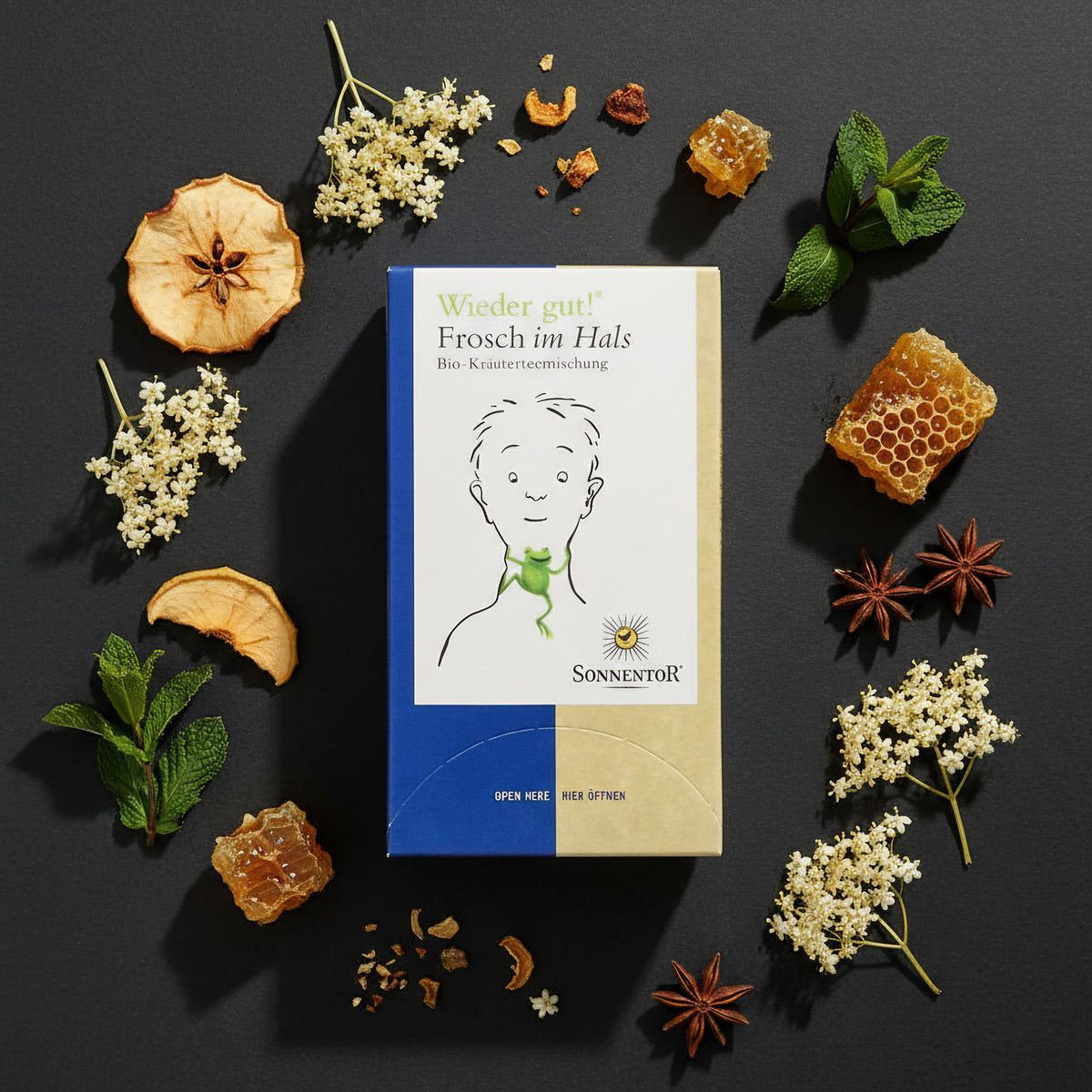 An artistic overhead shot of the 'Sonnentor Frosch im Hals Bio-Kräutertee' box with vibrant herbal ingredients like dried apple slices, honeycomb, and elderflowers artfully scattered around it on a matte, textured surface. HDR studio lighting with rim ligh