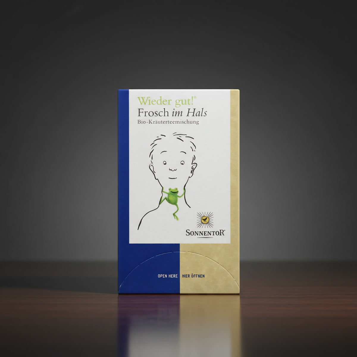 A close-up of the 'Sonnentor Frosch im Hals Bio-Kräutertee' package placed elegantly on a sleek, reflective dark wooden surface.