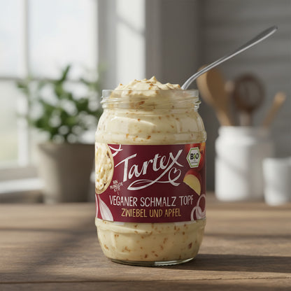 Close-up of Tartex Bio Freiburger Schmalz-Töpfle jar with open lid, showcasing creamy vegan spread with onion flecks and apple slices in a rustic kitchen.