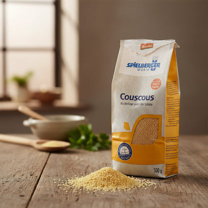 Close-up of Spielberger Bio Couscous grains spilling from a textured paper bag in a rustic kitchen, accented with herbs and wooden utensils.