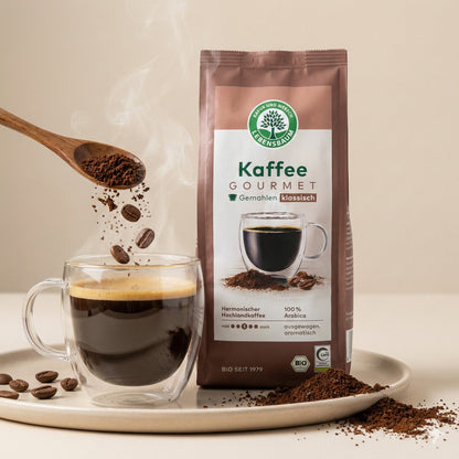 lebensbaum kaffee gourmet klassisch gemahlen neben dampfender Tasse Kaffee, Holzlöffel mit Kaffeepulver und Bohnen darüber