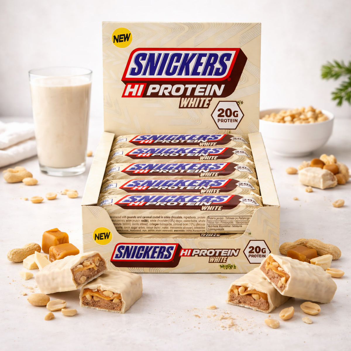 Snickers Hi-Protein White Bar 12x57g in geöffneter Verpackung, angeschnittene weiße schokoriegel im Vordergrund, im Hintergrund ein Glas Shake und Schale mit Erdnüssen.