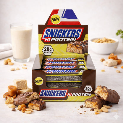 Snickers Protein Riegel 12x55g in geöffneter Verkaufspackung, umgeben von Erdnüssen, Karamell, Fudge und einem Glas Shake auf hellem Untergrund.