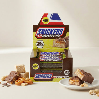 Snickers Eiweißriegel 12x55g in offener Box, angeschnittener Schokoriegel auf einem Teller, daneben Erdnüsse, Karamell und Fudge-Stücke.