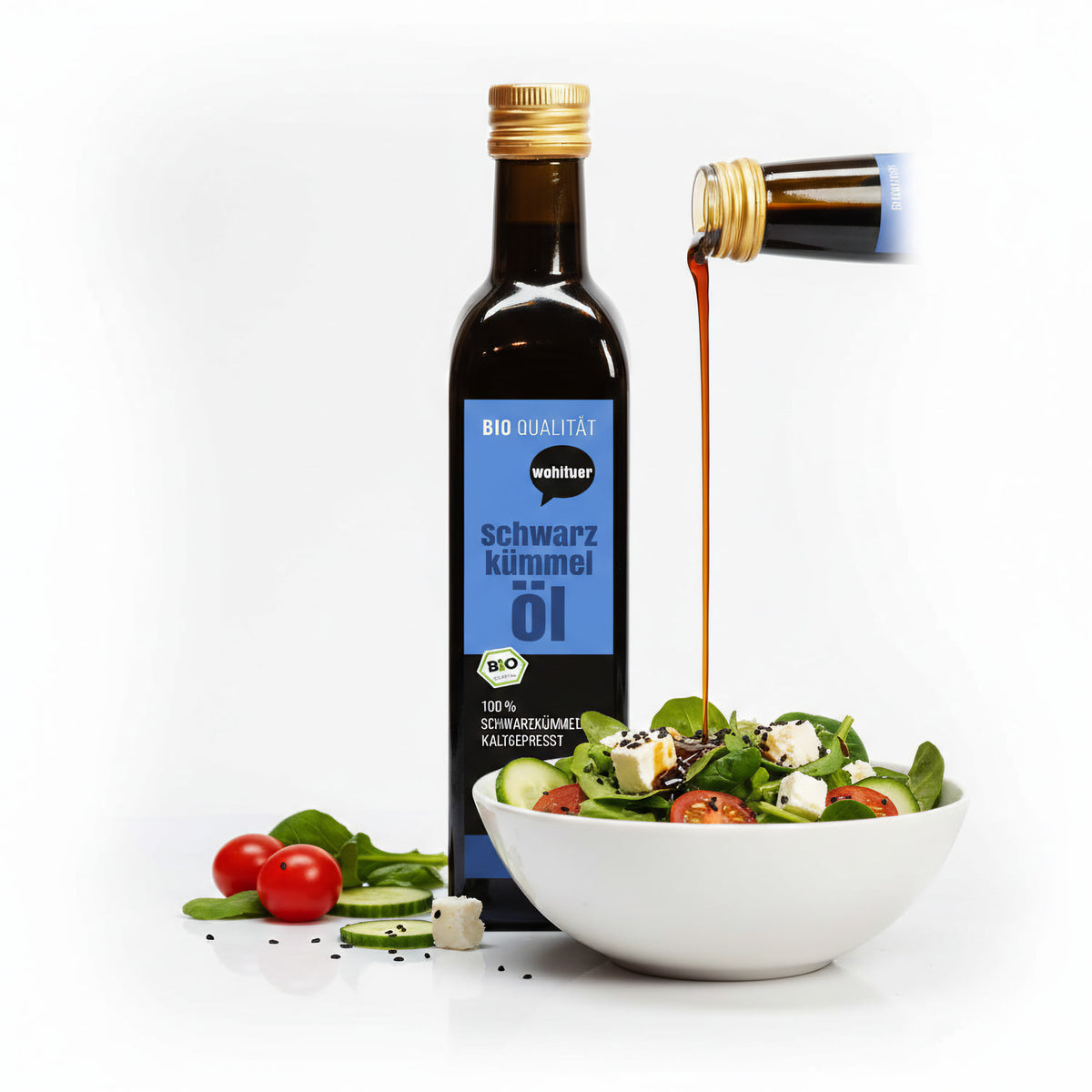 Eine Flasche wohltuer Schwarzkümmelöl kaltgepresst 250ml neben einer Schüssel Salat, Öl wird aus einer zweiten Flasche darüber gegossen.