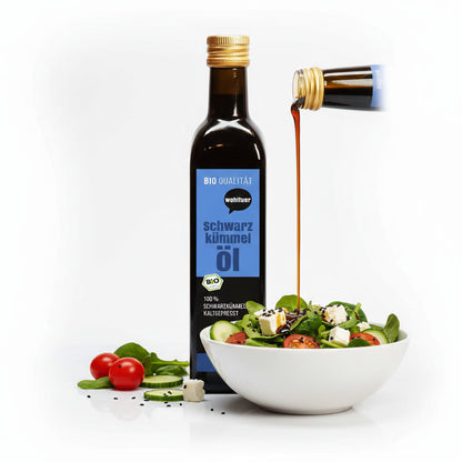 Eine Flasche wohltuer Schwarzkümmelöl kaltgepresst 250ml neben einer Schüssel Salat, Öl wird aus einer zweiten Flasche darüber gegossen.
