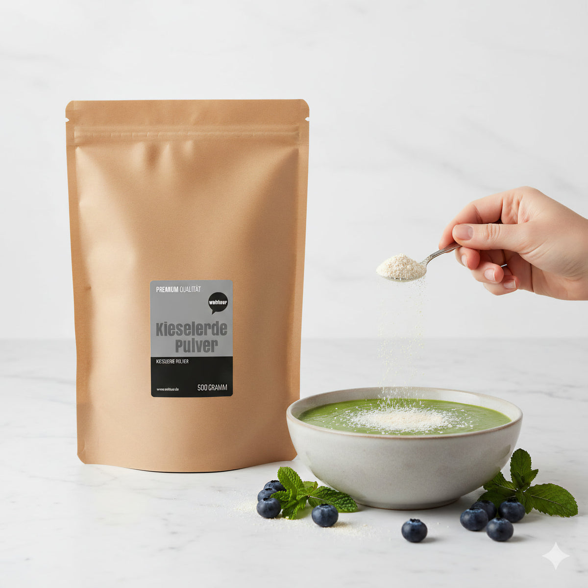 wohltuer Kieselerde Pulver 500g neben einer Schale mit grüner Flüssigkeit, eine Hand streut Pulver mit einem Löffel darüber, Blaubeeren und Minze im Vordergrund.
