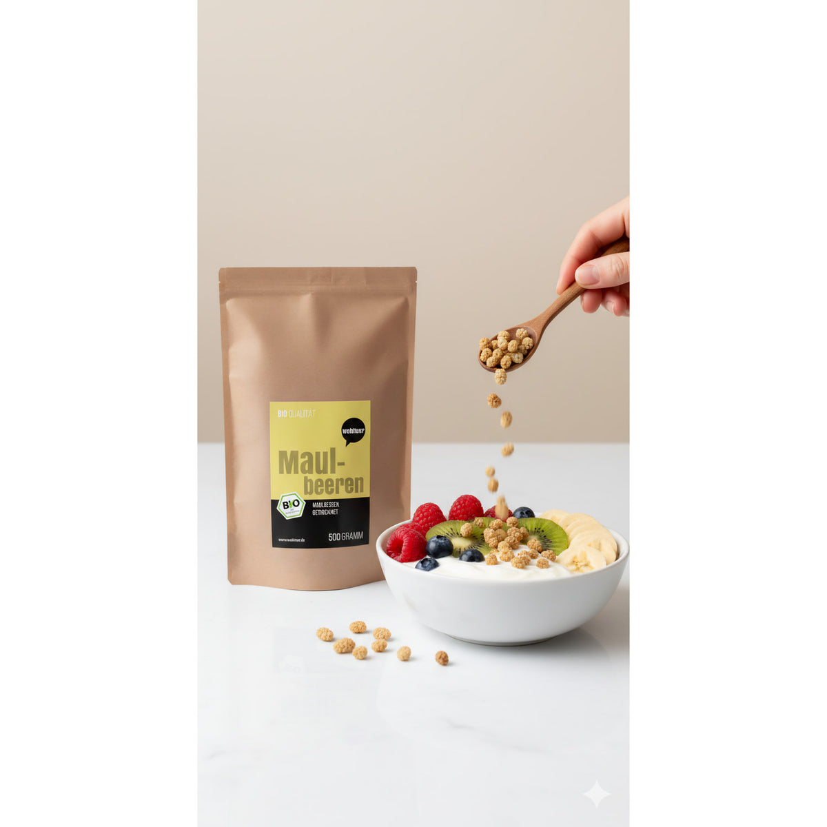 Beutel wohltuer Maulbeeren getrocknet 500g neben einer Frühstücksbowl mit frischen Früchten und einer Hand, die Maulbeeren mit einem Löffel darüber streut.