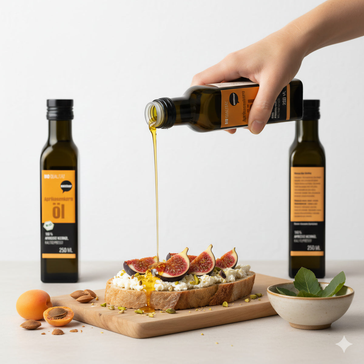 Hand gießt wohltuer Aprikosenkernöl kaltgepresst über ein mit Feigen belegtes Brot, daneben Aprikosen, Kerne und weitere Flaschen auf einem Tisch.