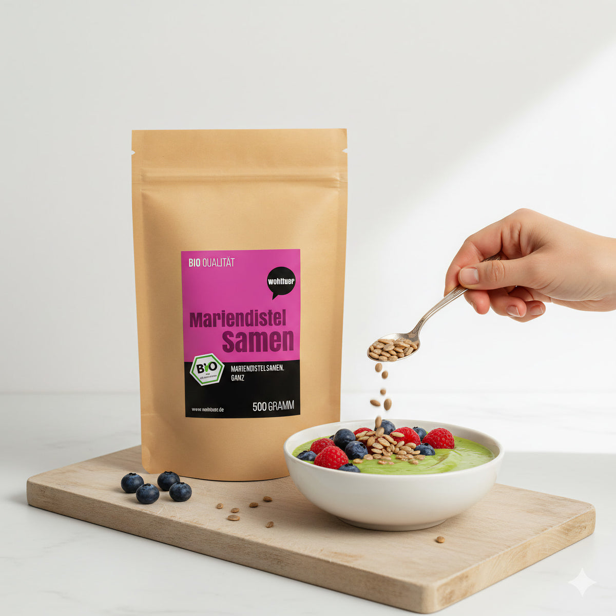 Wohltuer Mariendistelsamen ganz 500g auf Holzbrett, daneben Schale mit Smoothie und Beeren, Hand streut Samen mit Löffel darüber.