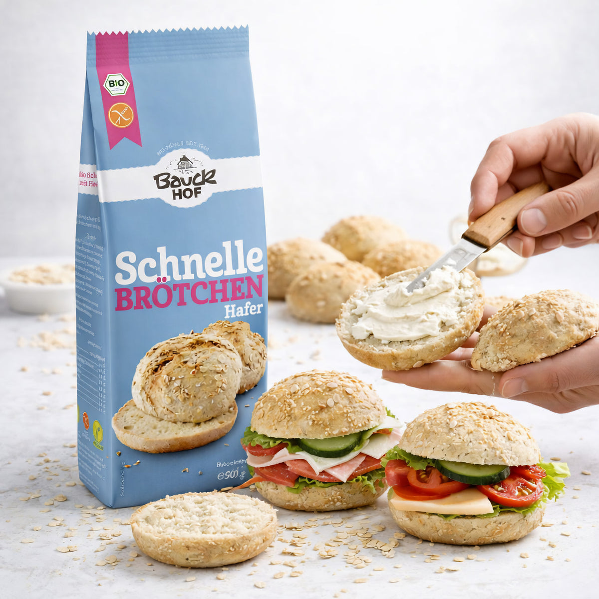 Bauckhof Backmischung Schnelle Brötchen Hafer, Packung neben belegten Brötchen und einer Hand, die ein Brötchen mit Aufstrich bestreicht.