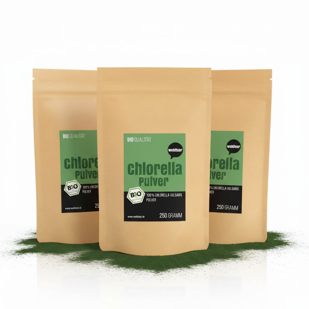 Drei braune Standbeutel wohltuer Chlorella Algenpulver 250g, stehend auf grünem Pulver, Vorderseiten mit grün-schwarzem Etikett sichtbar.