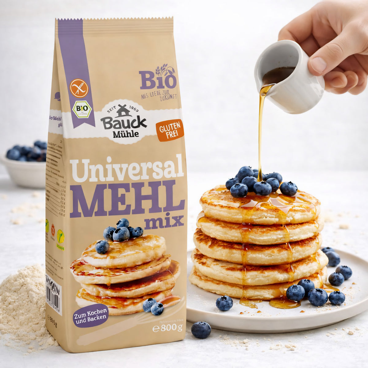 Bauckhof Universal Mehl-Mix 800g neben einem Stapel Pancakes mit Blaubeeren, über die Sirup gegossen wird, im Hintergrund eine Schale mit Blaubeeren.