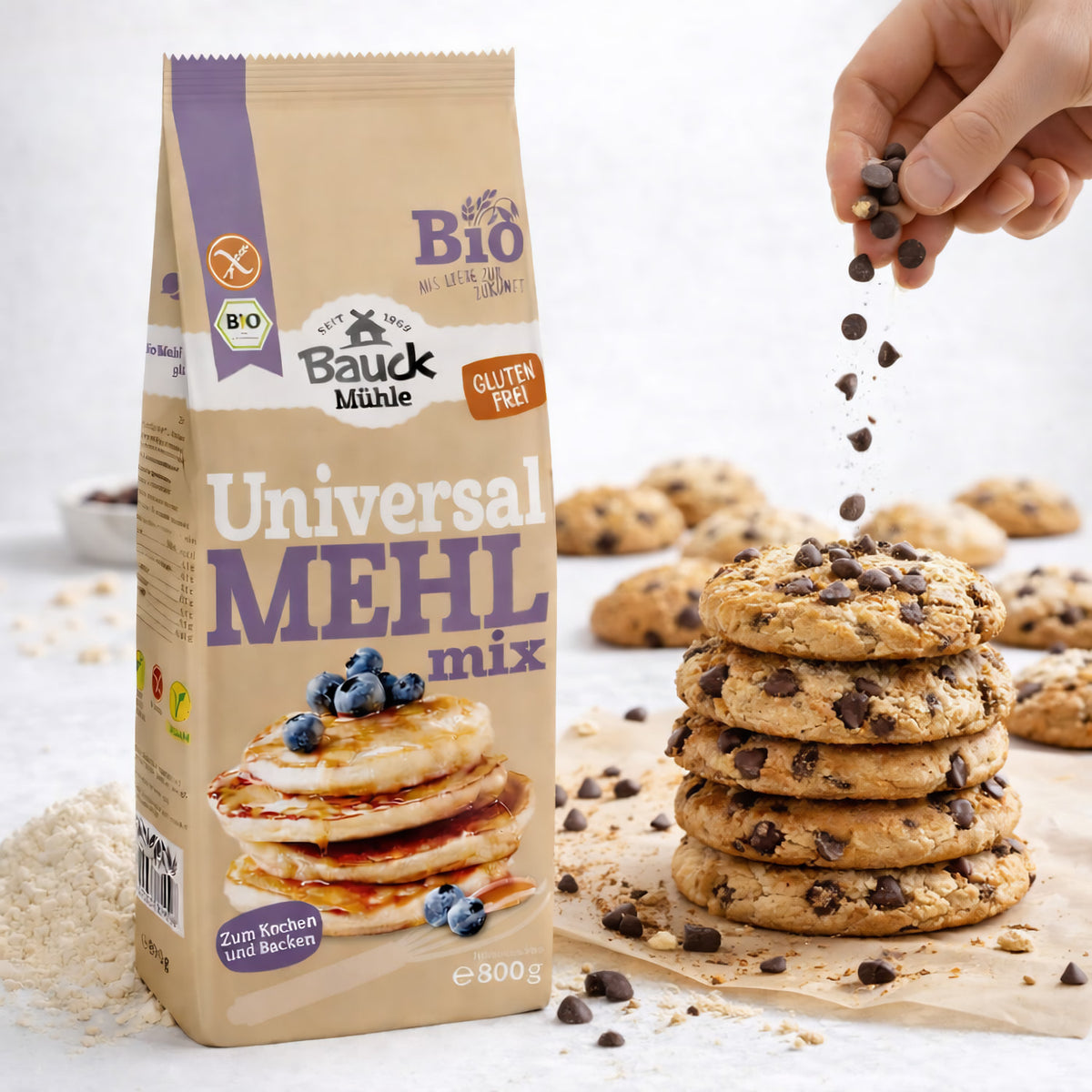 Bauckhof Mehl-Mix Universal glutenfrei 800g neben einem Stapel Cookies mit Schokostückchen, darüber werden weitere Schokodrops gestreut.