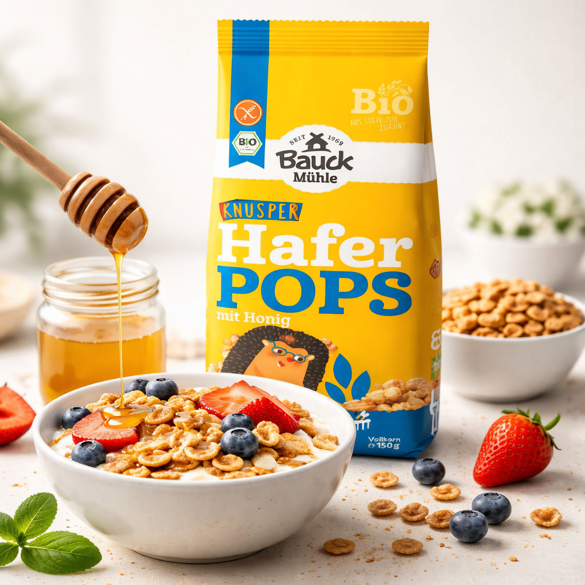 Bauckhof Haferpops mit Honig neben einer Schale Joghurt mit Pops, Erdbeeren und Blaubeeren, Honig wird darüber geträufelt, im Hintergrund weitere Frühstückszutaten.