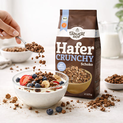 Bauckhof Hafer Crunchy Schoko neben einer Schale Joghurt mit frischen Beeren und Bananenscheiben, eine Hand streut crunchy Schoko darüber.