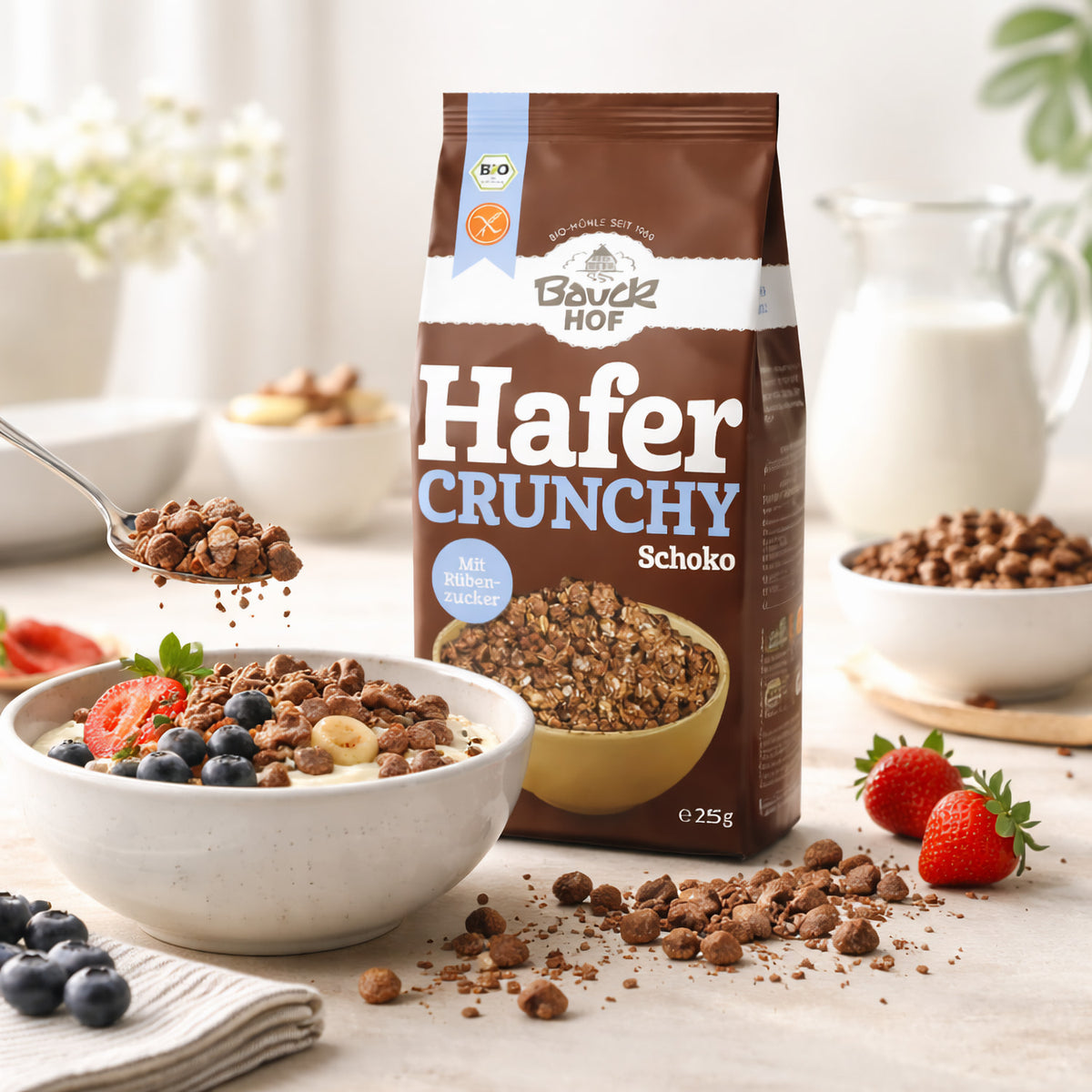 Schale mit Joghurt, Hafer Crunchy Schoko, Beeren und Bananen, daneben Bauckhof Hafer Crunchy Schoko Packung, im Hintergrund Milch und Erdbeeren.