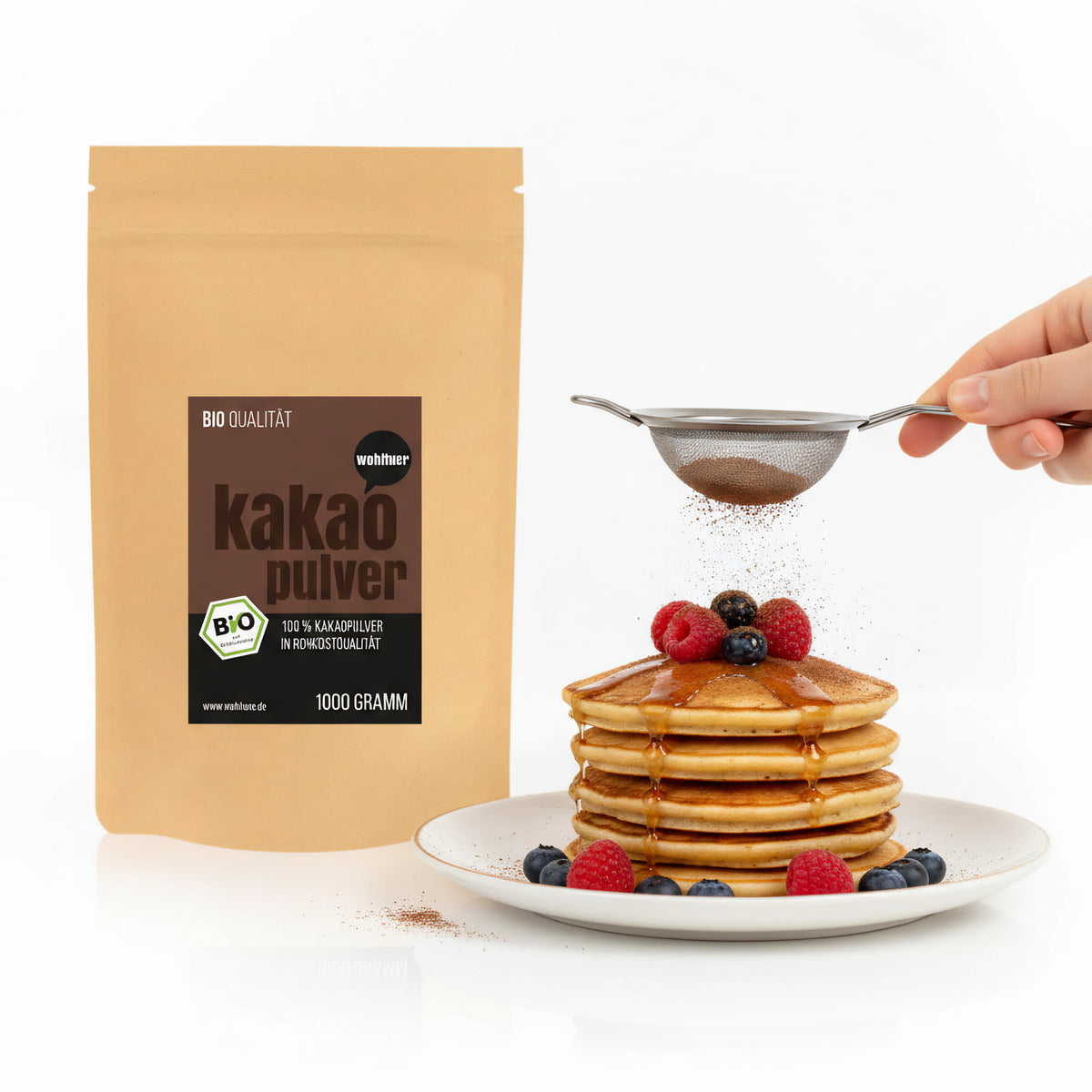 Beutel wohltuer Kakaopulver kaltgepresst 1000g neben einem Teller Pancakes mit Beeren, darüber wird Kakaopulver gesiebt.
