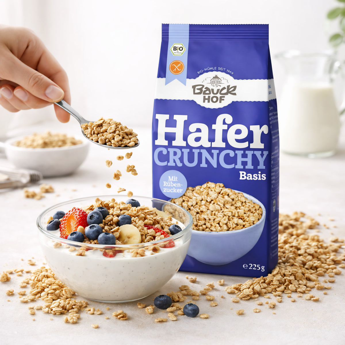 Schale mit Joghurt, frischen Beeren und Bauckhof Hafer Crunchy Basis, daneben eine Hand mit Löffel, im Hintergrund die Produktverpackung.