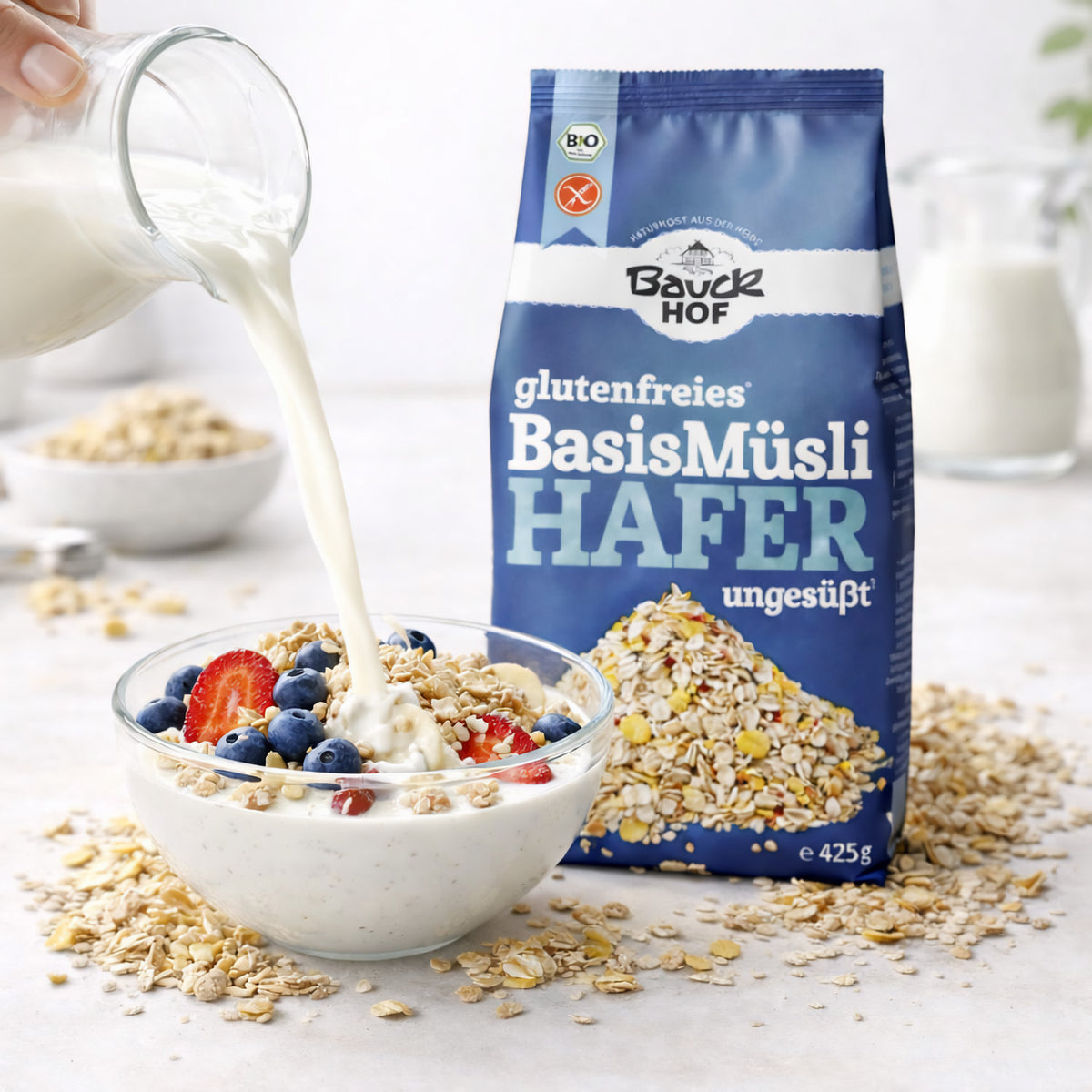 Milch wird in eine Schale mit Bauckhof Hafermüsli Basis glutenfrei, Joghurt und Beeren gegossen, Packung im Hintergrund.