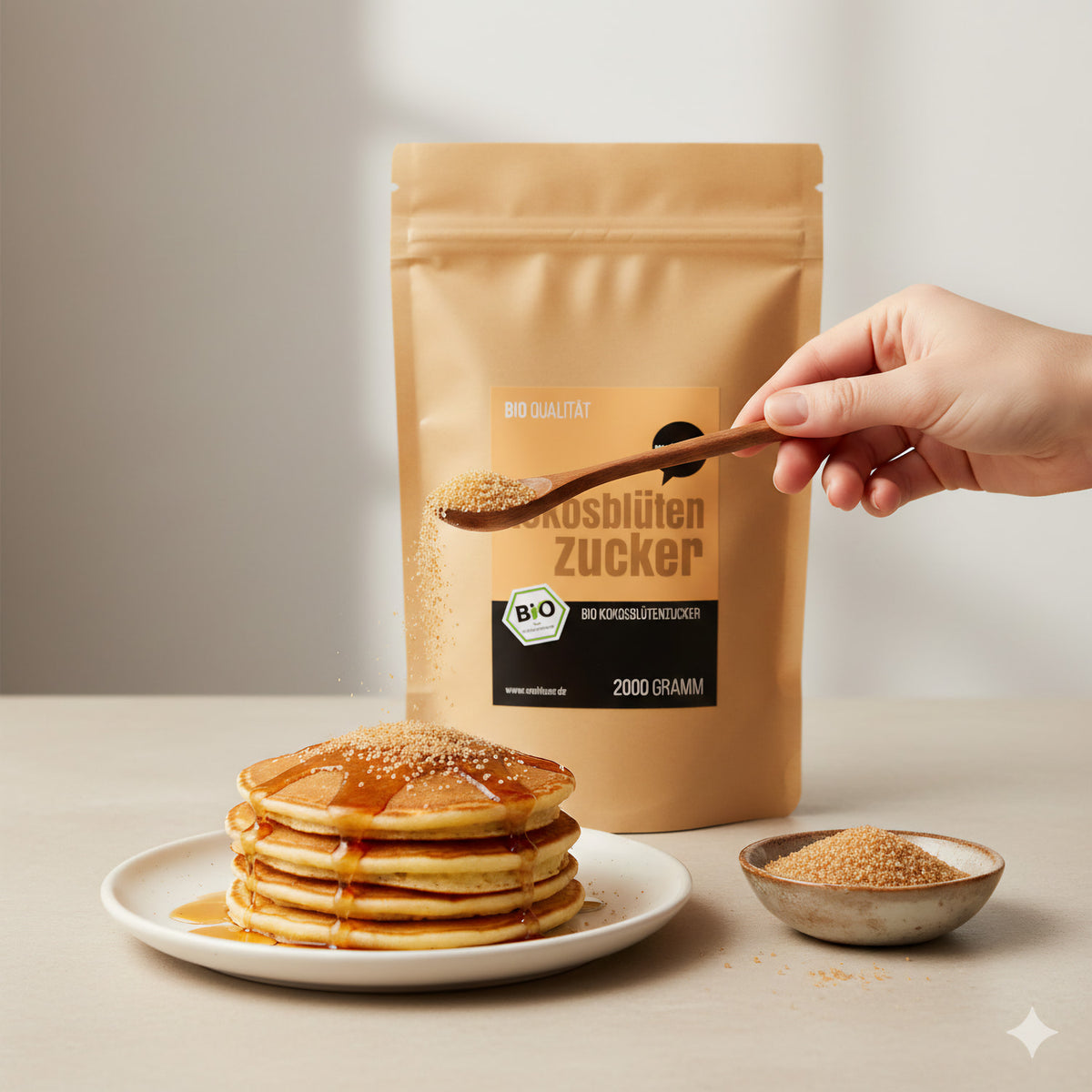 Hand streut wohltuer Kokosblütenzucker mit Löffel über Pancakes, Packung und Schale mit Zucker im Hintergrund.