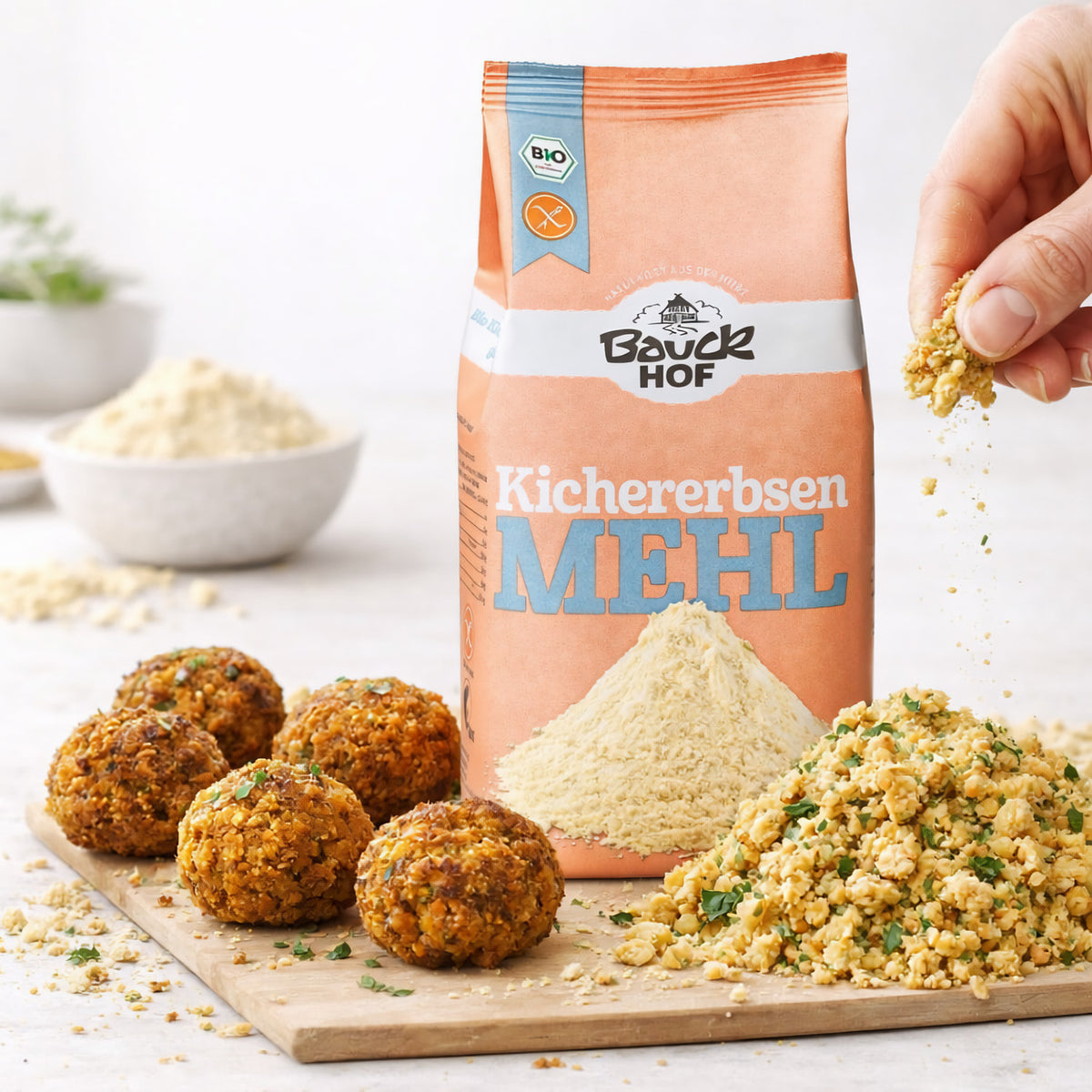 Kichererbsenmehl von Bauckhof auf einem Tisch, daneben Falafelbällchen und eine Hand, die gewürztes Kichererbsen-Superfood auf ein Holzbrett streut.