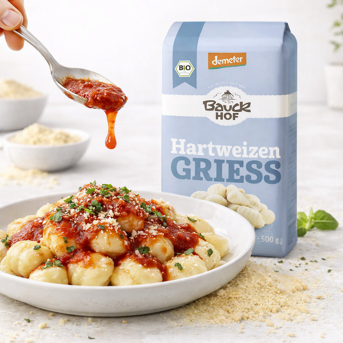 Bauckhof Hartweizengrieß neben einem Teller Gnocchi mit Tomatensauce, im Vordergrund wird Sauce mit einem Löffel über die Gnocchi gegeben.
