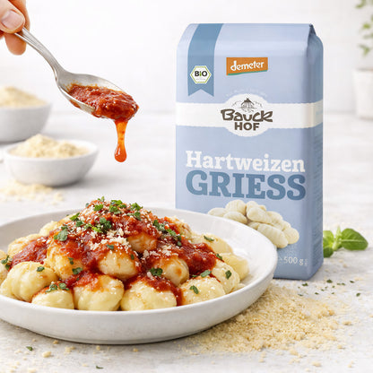 Bauckhof Hartweizengrieß neben einem Teller Gnocchi mit Tomatensauce, im Vordergrund wird Sauce mit einem Löffel über die Gnocchi gegeben.
