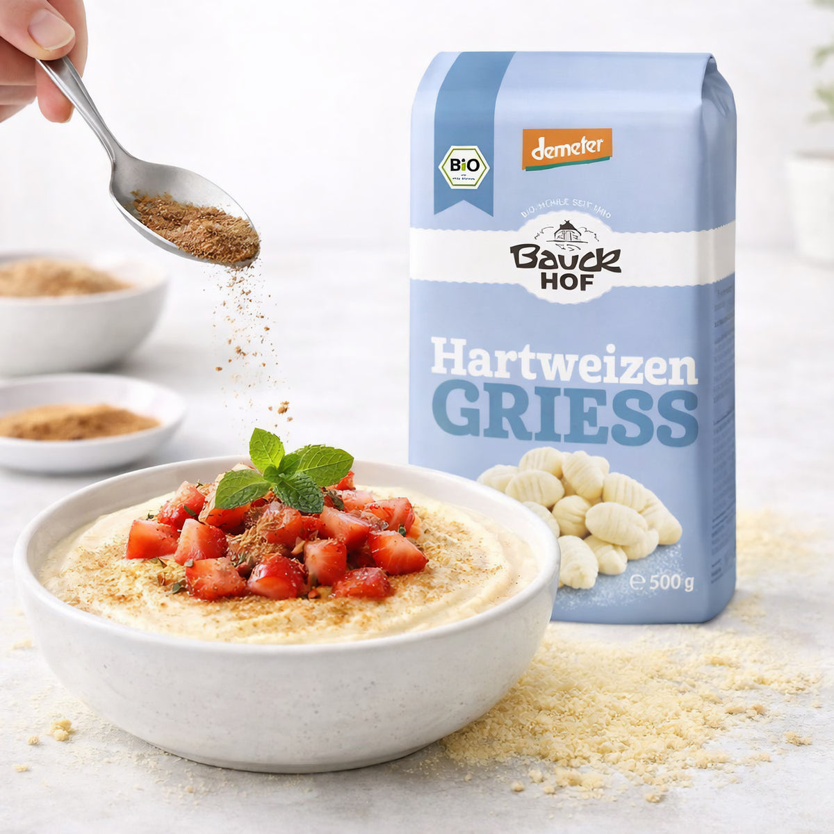 Bauckhof Hartweizengrieß neben einer Schale Grießbrei mit Erdbeeren und Minze, darüber wird Zimt mit einem Löffel gestreut.