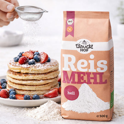 Bauckhof Reismehl hell glutenfrei neben einem Stapel Pancakes mit Beeren, im Vordergrund etwas loses Mehl, im Hintergrund wird Puderzucker gestreut.