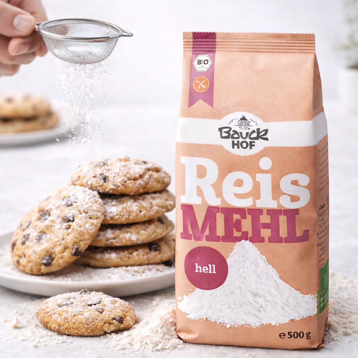 Bauckhof Reismehl hell glutenfrei neben einem Teller mit gestapelten Cookies, im Vordergrund loses Mehl, darüber wird Puderzucker gesiebt.
