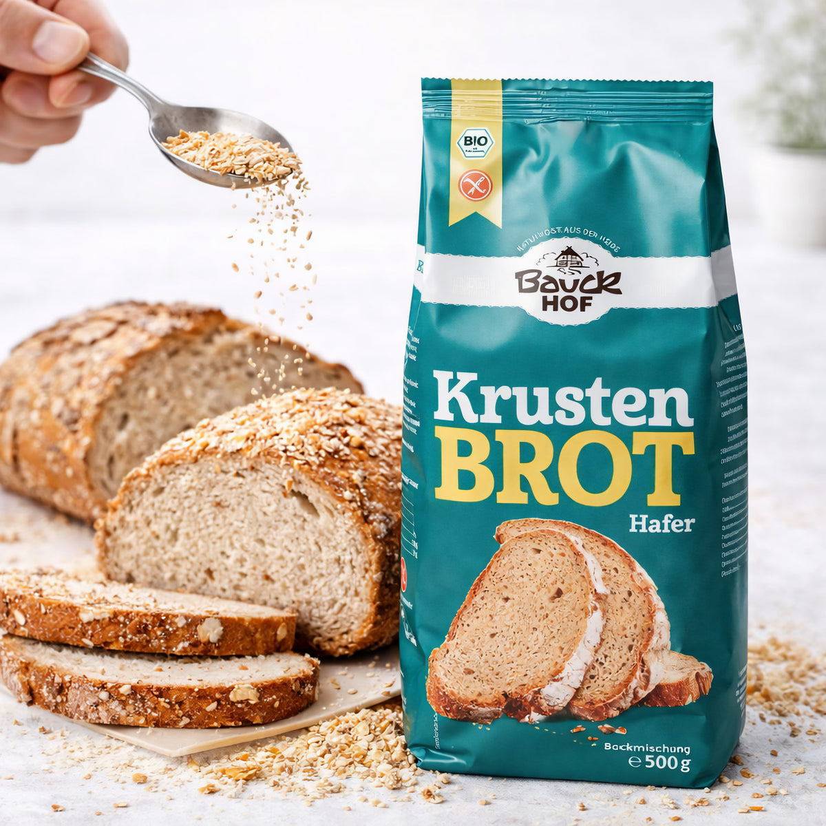 bauckhof krustenbrot glutenfrei 500g neben aufgeschnittenem brot, darüber werden mit einem löffel körner gestreut.