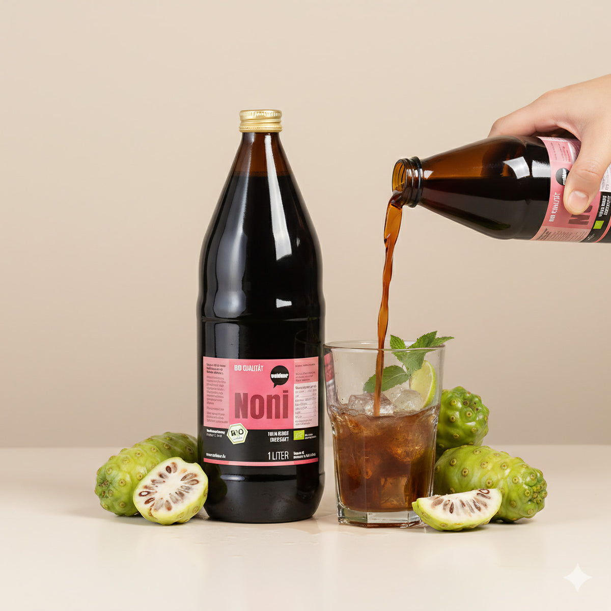 Wohltuer Noni Direktsaft wird aus einer Flasche in ein Glas mit Eis, Limette und Minze gegossen, daneben liegen Noni-Früchte.