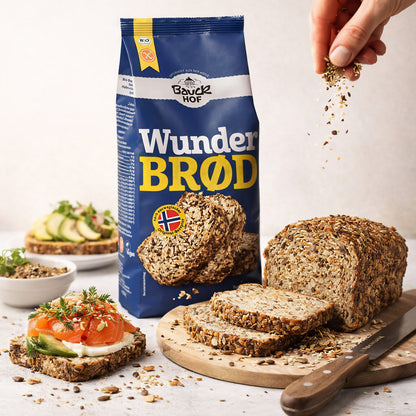 Bauckhof Wunderbrot Backmischung 600g neben angeschnittenem Brotlaib und belegter Brotscheibe, Hand streut Körner darüber.