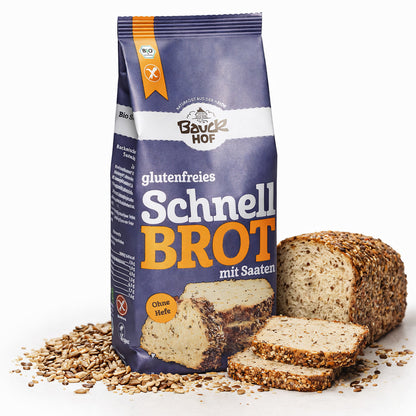 bauckhof schnellbrot backmischung glutenfrei 500g neben einem angeschnittenen brotlaib mit saatkruste und brotscheiben, umgeben von körnern auf weißem untergrund.