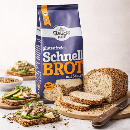 bauckhof schnellbrot backmischung glutenfrei 500g mit angeschnittenem brotlaib und brotscheiben auf einem holzbrett, daneben belegte brotscheiben und eine schale mit saatmischung.