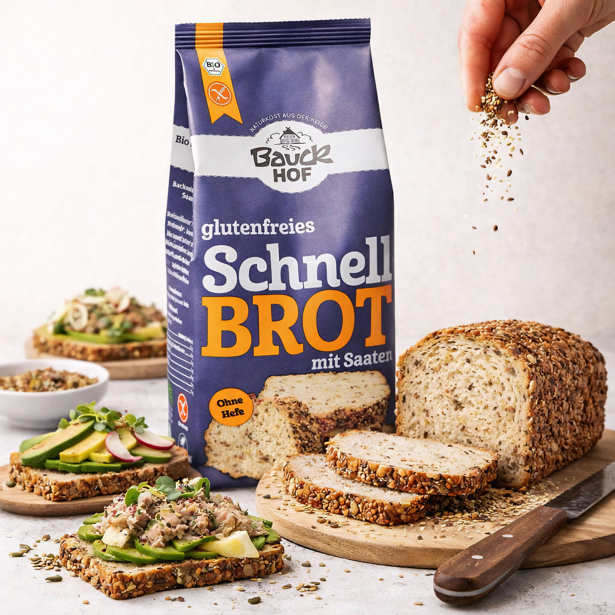 bauckhof schnellbrot backmischung glutenfrei 500g mit brotlaib und brotscheiben auf einem brett, im vordergrund eine hand, die saatmischung über das brot streut, dazu belegte brotscheiben.
