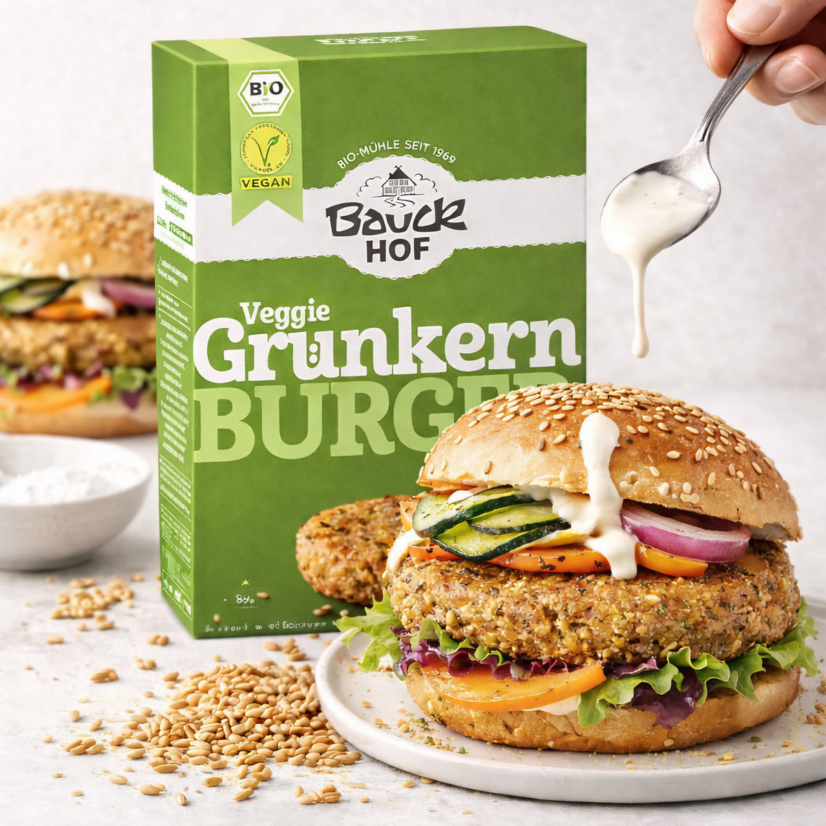 Vegane Burger mit Grünkern-Pattie von Bauckhof im Sesambrötchen, darüber wird mit einem Löffel Sauce gegeben, Packung im Hintergrund.