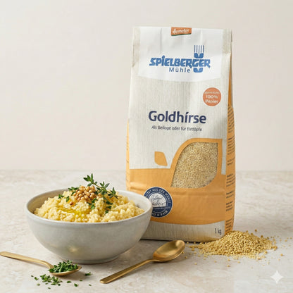 Packung Spielberger Goldhirse neben einer Schale gekochter Hirse mit Kräutern und goldfarbenem Löffel auf hellem Tisch.