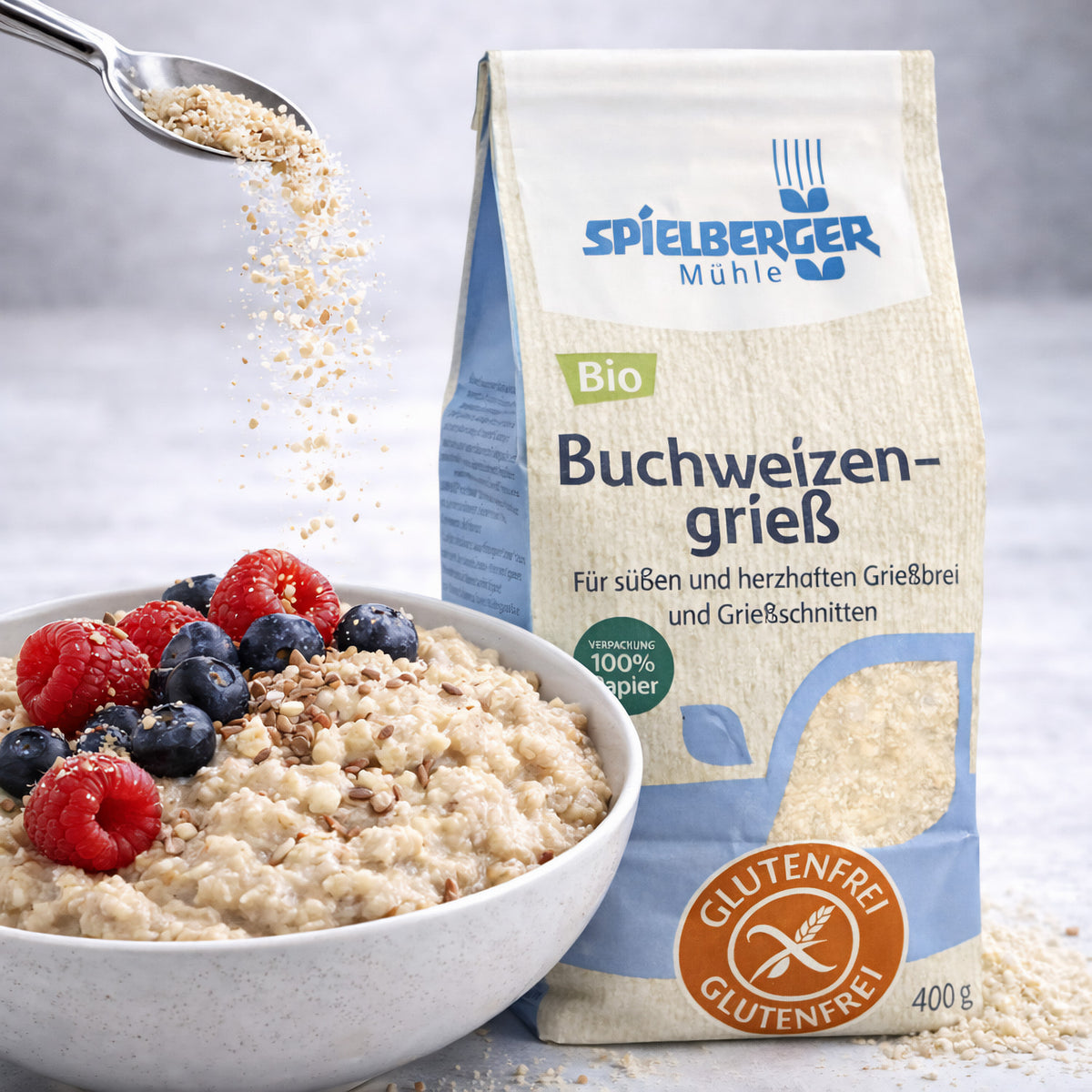 spielberger buchweizengrieß 400g neben einer Schale Frühstücksbrei mit Beeren, ein Löffel streut Grieß darüber.