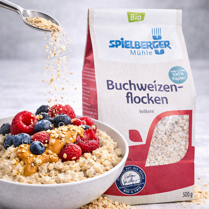 schale mit buchweizenflocken von spielberger, garniert mit frischen Beeren und Nussmus, daneben die 500g-Packung, Flocken werden mit einem Löffel darüber gestreut