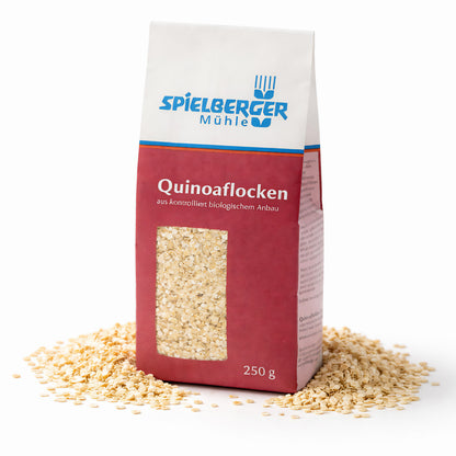 Packung Spielberger Quinoaflocken 250g steht aufrecht, umgeben von verstreuten Quinoaflocken auf weißem Hintergrund.