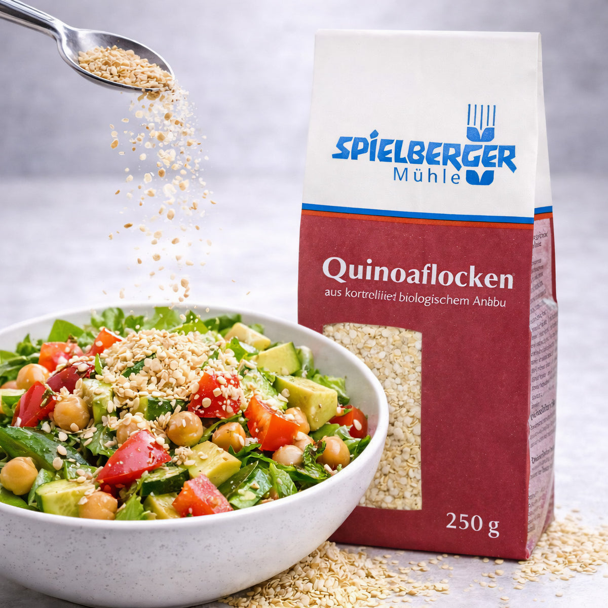 Spielberger Quinoaflocken 250g neben einer Schüssel Salat, Quinoaflocken werden mit einem Löffel über den Salat gestreut.
