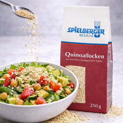 Spielberger Quinoaflocken 250g neben einer Schüssel Salat, Quinoaflocken werden mit einem Löffel über den Salat gestreut.